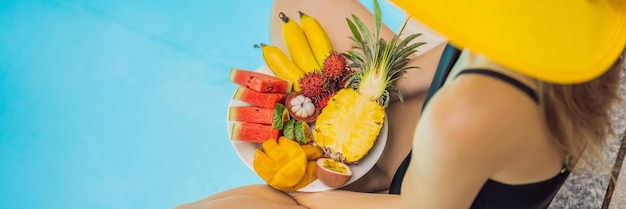 Giovane donna che si rilassa e mangia un piatto di frutta vicino alla piscina dell'hotel Dieta esotica estiva Foto di gambe con cibo sano vicino alla piscina vista dall'alto Stile di vita della spiaggia tropicale BANNER LONG FORMAT