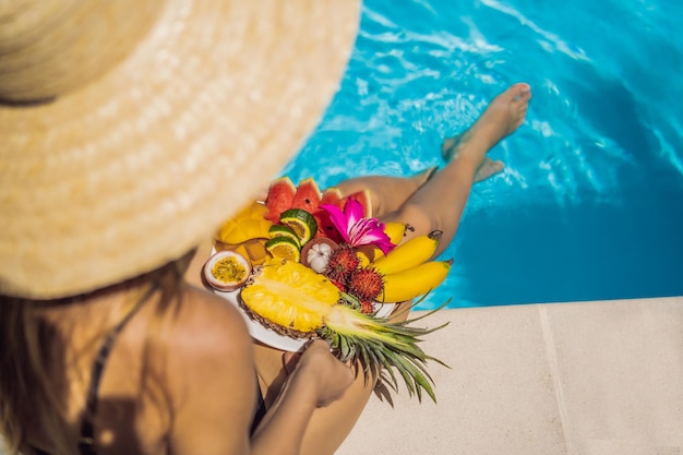 Giovane donna che si rilassa e mangia un piatto di frutta vicino alla piscina dell'hotel Dieta esotica estiva Foto di gambe con cibo sano vicino alla piscina vista dall'alto Stile di vita della spiaggia tropicale