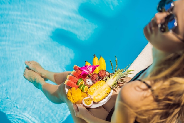 Giovane donna che si rilassa e mangia un piatto di frutta vicino alla piscina dell'hotel Dieta esotica estiva Foto di gambe con cibo sano vicino alla piscina vista dall'alto Stile di vita della spiaggia tropicale