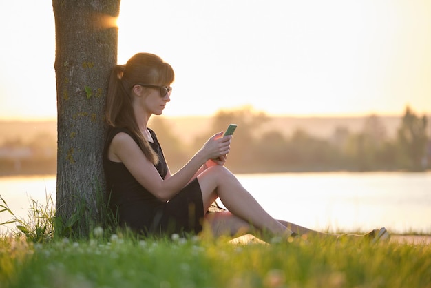 Giovane donna che si rilassa sul prato in riva al lago navigando con il suo telefono cellulare all'aperto in una calda serata estiva Concetto di comunicazione e connessione mobile
