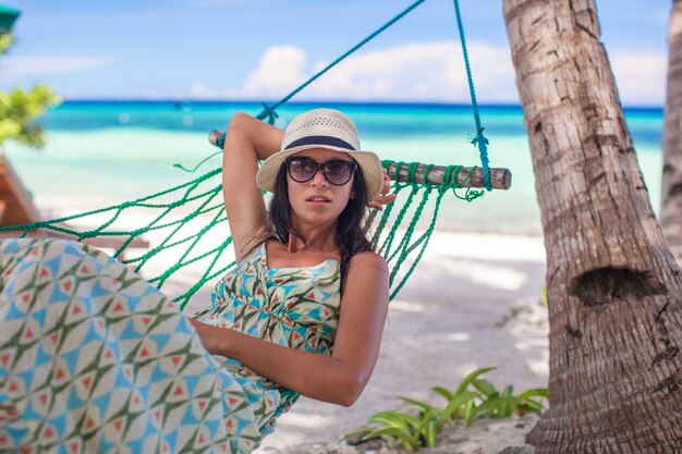 Giovane donna che si siede all'amaca nell'ombra dell'albero su una spiaggia