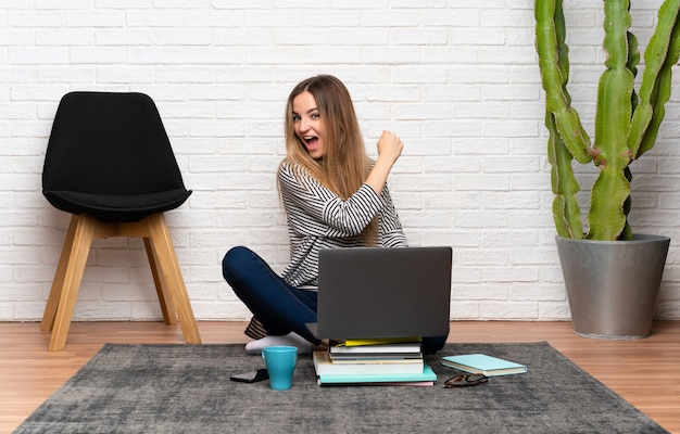 Giovane donna che si siede sul pavimento con il suo computer portatile che celebra una vittoria