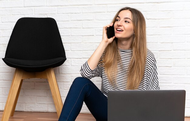 Giovane donna che si siede sul pavimento con il suo portatile parlando con cellulare