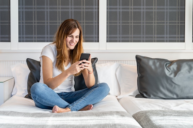 Giovane donna che si siede sul suo letto usando il suo telefono cellulare