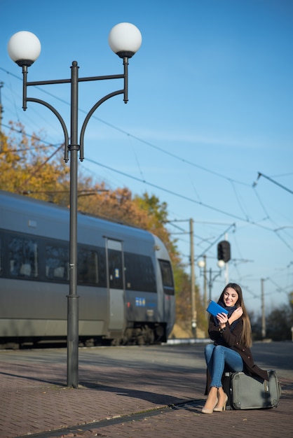 Giovane donna che si siede sulla valigia facendo uso di una compressa alla stazione ferroviaria