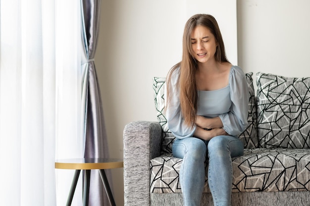 Giovane donna che soffre di forte mal di stomaco mentre è seduta sul divano di casa. problemi di salute