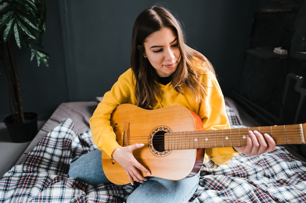 Giovane donna che suona la chitarra acustica a casa, seduta sul letto