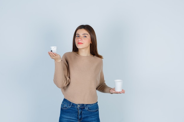 Giovane donna che tiene bicchieri di carta in maglione, jeans e guardando malinconico, vista frontale.