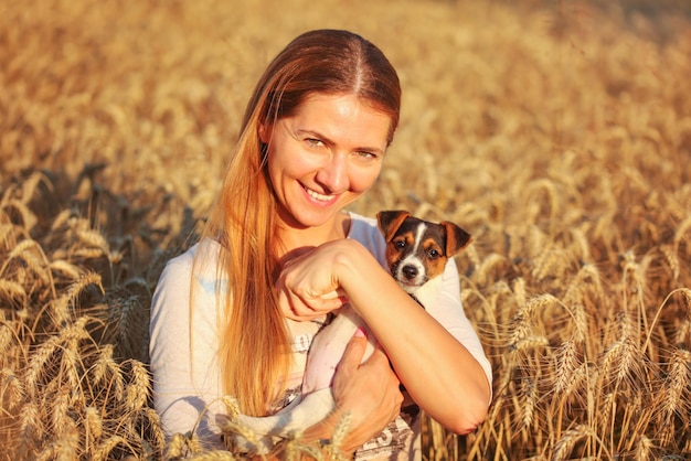 Giovane donna che tiene il cucciolo di Jack Russell terrier sulle sue mani, entrambi sorridenti, campo di grano illuminato al tramonto sullo sfondo.