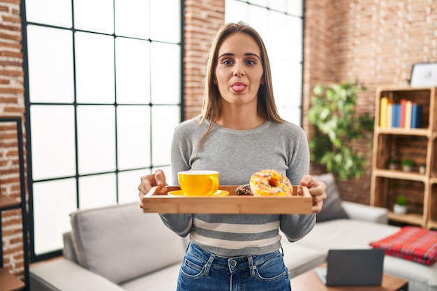 Giovane donna che tiene il vassoio con il cibo per la colazione che attacca la lingua fuori felice con l'espressione divertente.