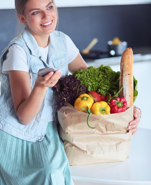 Giovane donna che tiene la borsa della spesa con verdure in piedi in cucina