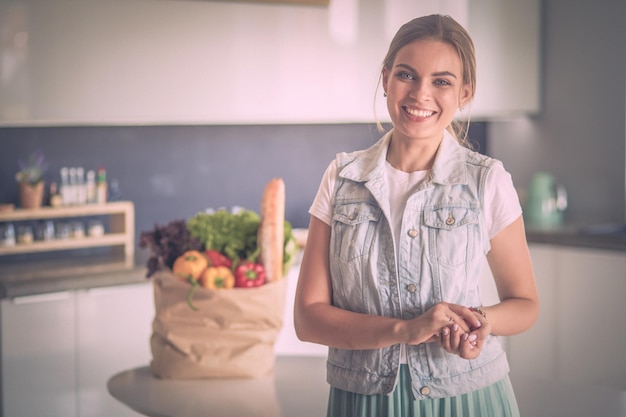 Giovane donna che tiene la borsa della spesa con verdure in piedi in cucina