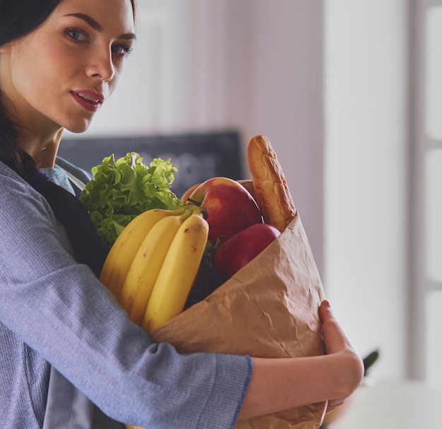 Giovane donna che tiene la borsa della spesa con verdure in piedi in cucina
