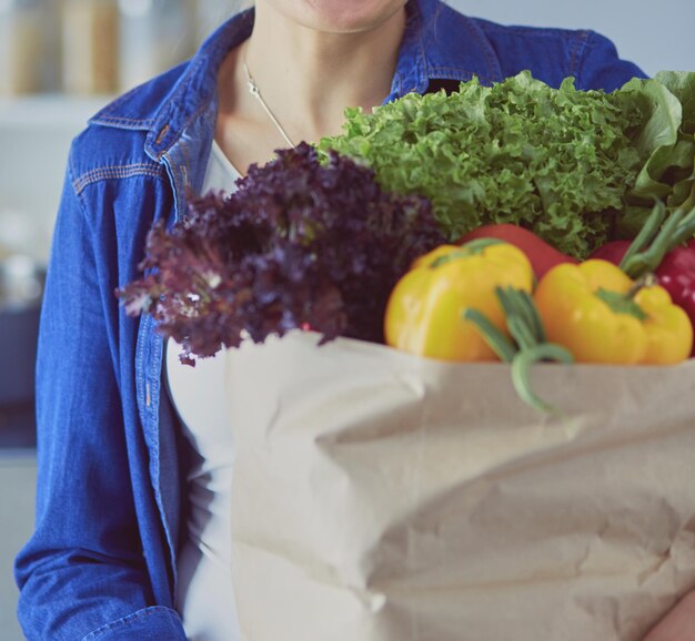 Giovane donna che tiene la borsa della spesa con verdure in piedi in cucina