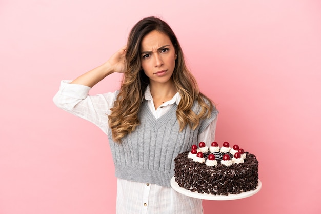 Giovane donna che tiene la torta di compleanno sopra fondo rosa isolato che ha dubbi