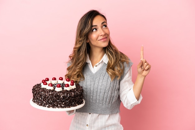 Giovane donna che tiene la torta di compleanno sopra fondo rosa isolato che indica una grande idea