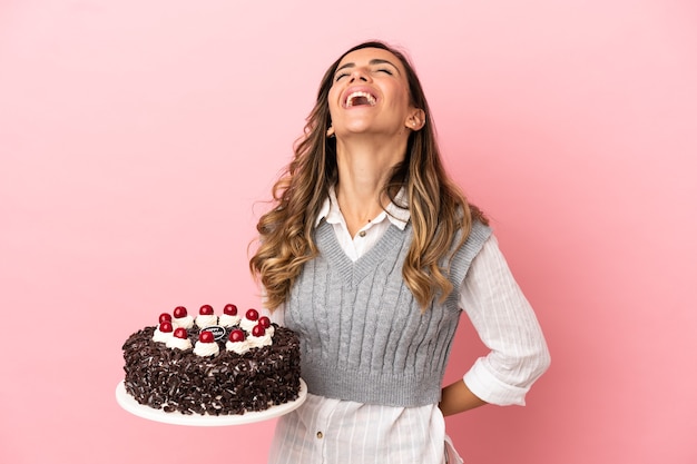 Giovane donna che tiene la torta di compleanno su sfondo rosa isolato ridendo