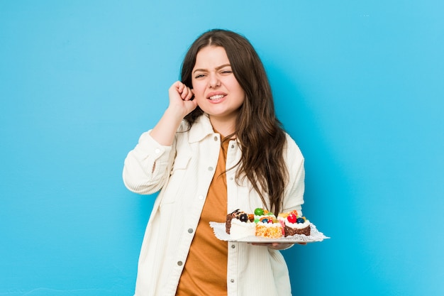 Giovane donna che tiene le torte dolci che coprono le orecchie con le mani.