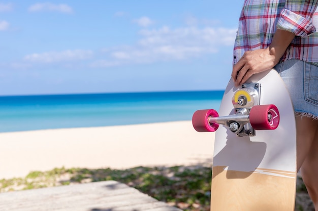 Giovane donna che tiene lo skateboard della vecchia scuola e in piedi accanto a una spiaggia