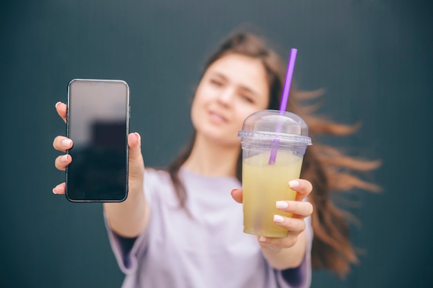 Giovane donna che tiene smartphone nero e tazza di limonata nelle mani e mostrandolo