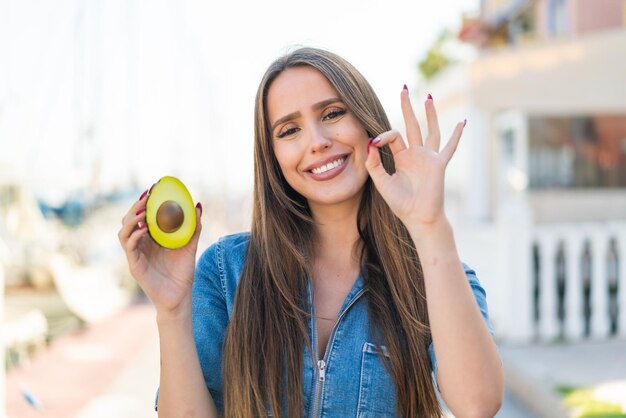Giovane donna che tiene un avocado all'aperto mostrando il segno ok con le dita