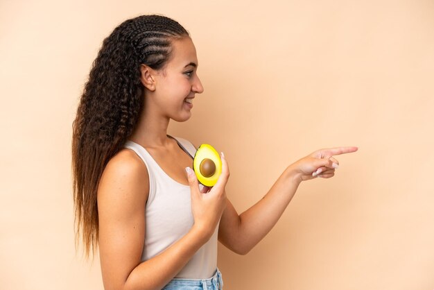 Giovane donna che tiene un avocado isolato su sfondo beige che indica il lato per presentare un prodotto