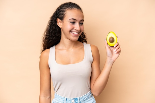 Giovane donna che tiene un avocado isolato su sfondo beige con felice espressione