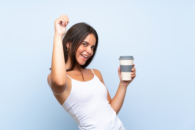 Giovane donna che tiene un caffè da asporto sopra la parete blu isolata che celebra una vittoria