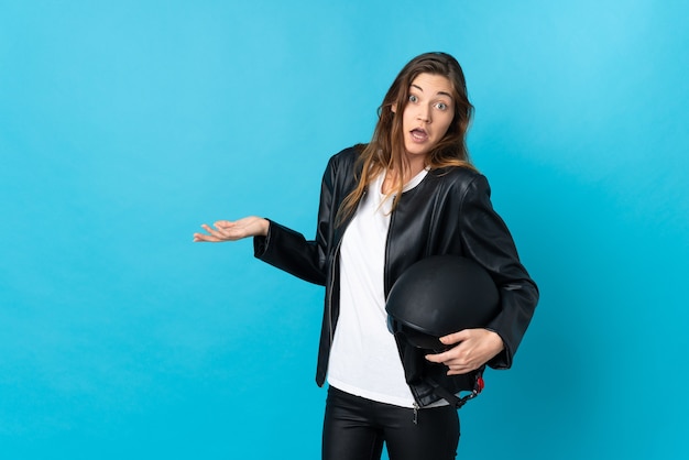 Giovane donna che tiene un casco da motociclista isolato sulla parete blu con espressione di sorpresa mentre guarda di lato