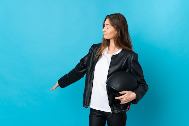 Giovane donna che tiene un casco del motociclo isolato sulla parete blu che estende le mani a lato per invitare a venire