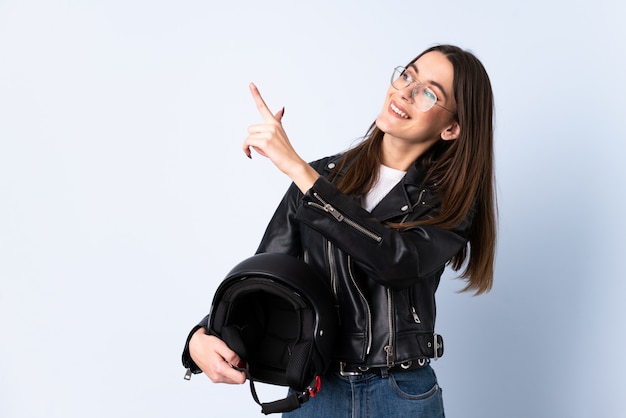 Giovane donna che tiene un casco del motociclo sopra la parete blu isolata che indica con il dito indice una grande idea