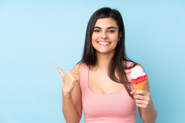 Giovane donna che tiene un gelato alla cornetta sopra la parete blu isolata che indica il lato per presentare un prodotto