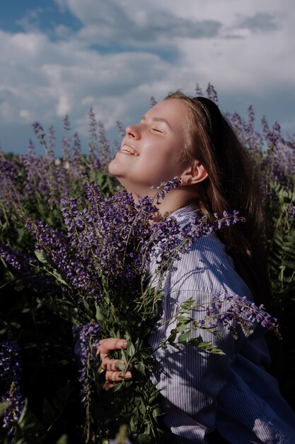Giovane donna che tiene un mazzo di fiori viola in piedi tra un campo di fiori di campo davanti a sky