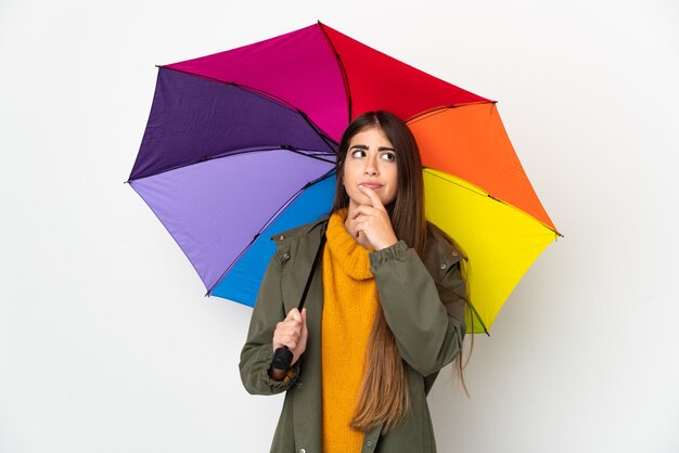 Giovane donna che tiene un ombrello isolato su priorità bassa bianca che ha dubbi mentre osserva in su