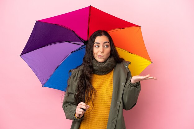 Giovane donna che tiene un ombrello isolato su sfondo rosa avendo dubbi mentre si alzano le mani