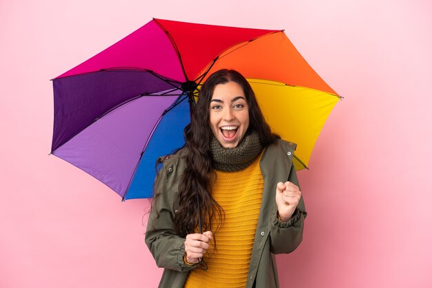 Giovane donna che tiene un ombrello isolato su sfondo rosa che celebra una vittoria nella posizione del vincitore