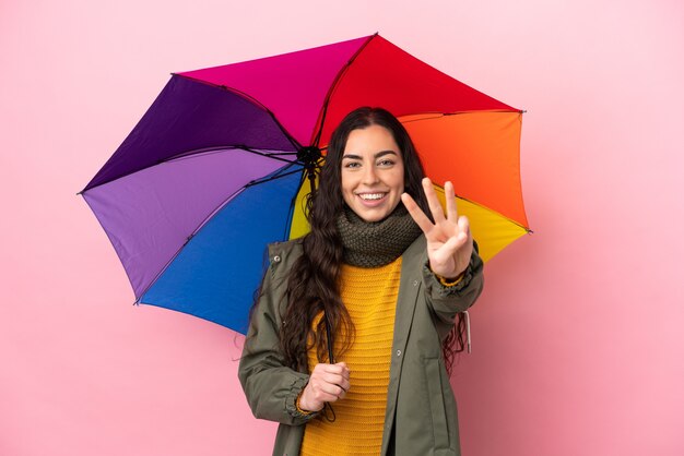 Giovane donna che tiene un ombrello isolato su sfondo rosa felice e contando tre con le dita