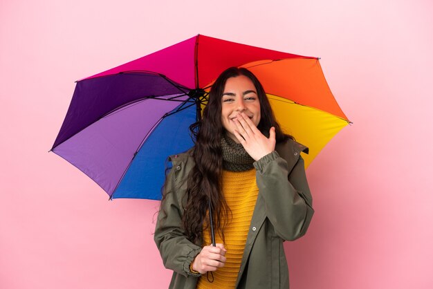 Giovane donna che tiene un ombrello isolato su sfondo rosa felice e sorridente che copre la bocca con la mano