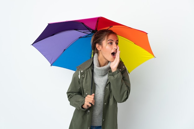 Giovane donna che tiene un ombrello isolato sul muro bianco che grida con la bocca spalancata di lato