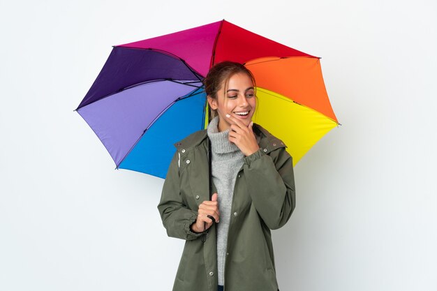 Giovane donna che tiene un ombrello isolato sul muro bianco che guarda al lato e sorridente