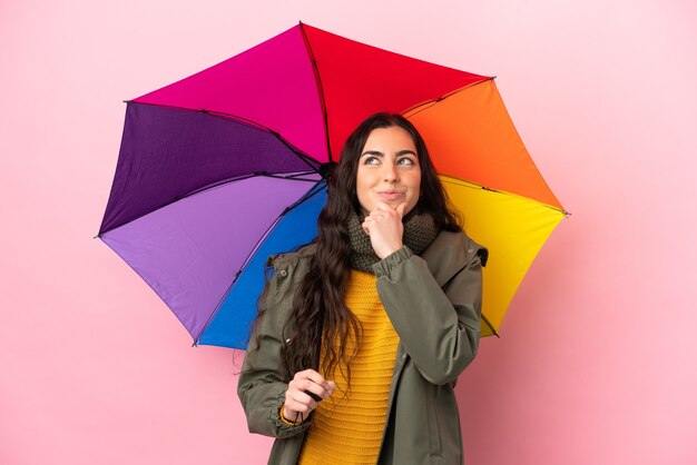 Giovane donna che tiene un ombrello isolato sul muro rosa e alzando lo sguardo
