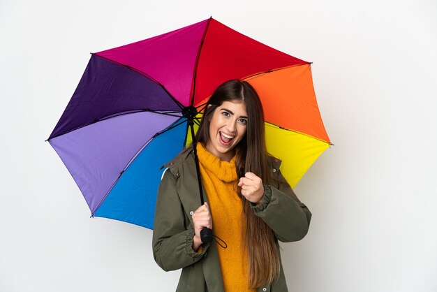 Giovane donna che tiene un ombrello isolato sulla parete bianca che celebra una vittoria
