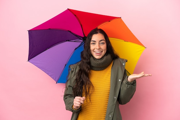 Giovane donna che tiene un ombrello isolato sulla parete rosa con l'espressione facciale scioccata