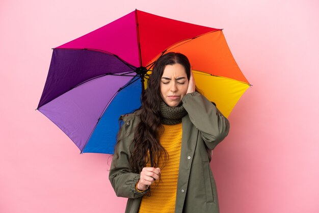 Giovane donna che tiene un ombrello isolato sulla parete rosa frustrata e che copre le orecchie