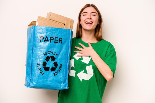 Giovane donna che tiene un sacchetto di riciclaggio pieno di carta da riciclare isolato su sfondo bianco ride ad alta voce tenendo la mano sul petto