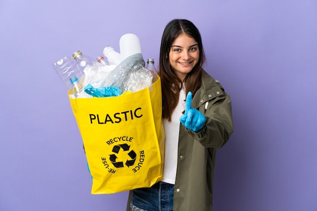 Giovane donna che tiene un sacchetto pieno di bottiglie di plastica da riciclare isolato sulla parete viola che mostra e alzando un dito