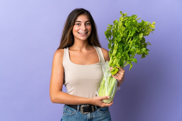 Giovane donna che tiene un sedano isolato sulla parete viola che sorride molto