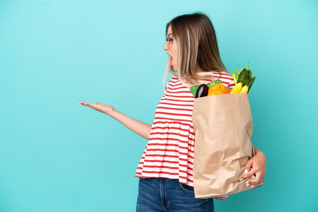 Giovane donna che tiene una borsa della spesa isolata su sfondo blu con espressione di sorpresa mentre guarda di lato