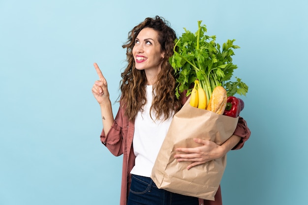 Giovane donna che tiene una borsa della spesa isolata sulla parete blu che indica una grande idea