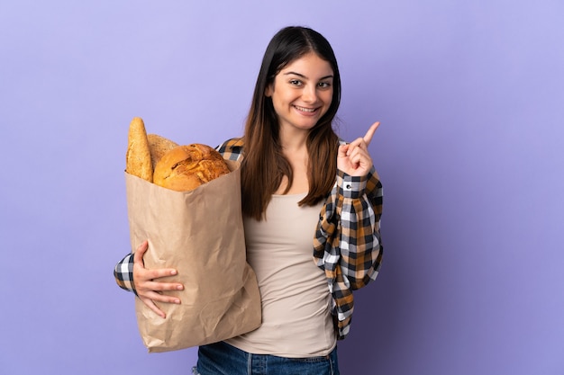 Giovane donna che tiene una borsa piena di pane isolato su viola rivolta verso l'alto una grande idea